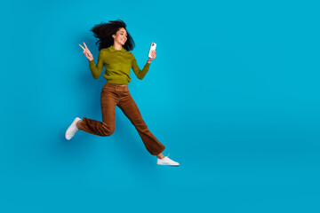 Obraz premium Energetic woman making peace sign while holding a smartphone against a blue background in casual stylish attire