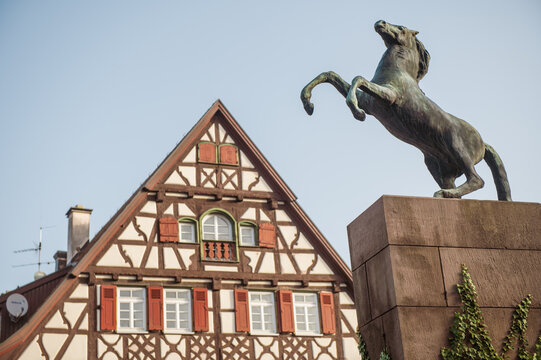Bronze-Skulptur  eines aufsteigenden Pferdes vor Giebelseite eines Fachwerkhauses mit Fensterladen und Sprossenfenstern vor blauem Himmel am Rossmarkt in Kirchheim unter Teck