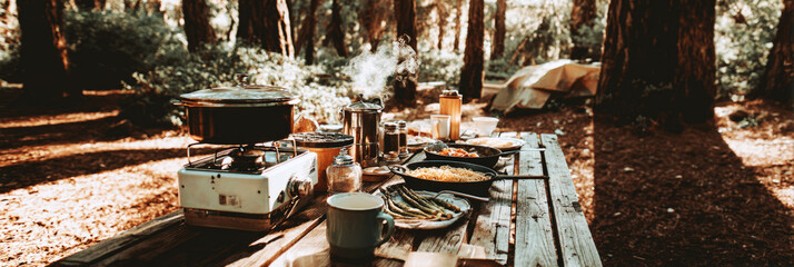 Campers prepare a meal outdoors using a portable stove, a kettle, and fresh ingredients, enjoying the tranquil forest environment in the background, banner