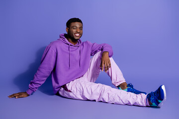 Young man in purple hoodie and pastel pants sitting and smiling on a purple background, showcasing modern casual style