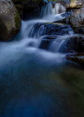 A small, natural waterfall flows between large rocks with a clear stream and a calming green atmosphere amidst a tropical forest