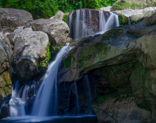 A small, natural waterfall flows between large rocks with a clear stream and a calming green...