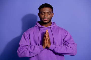Calm young man meditating with hands together, wearing purple hoodie, against a vibrant violet...