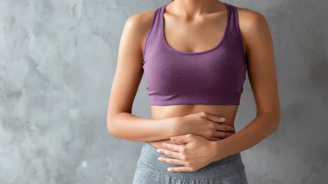 Fit athletic female in dark sportswear standing against textured gray wall, holding stomach area and experiencing potential menstrual discomfort or abdominal pain during wellness moment