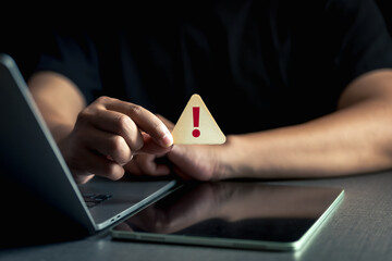 Cybersecurity alert concept. A person holds a warning sign with an exclamation mark near a laptop...
