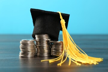 Scholarship. Graduate hat and coins on wooden table against light blue background, closeup