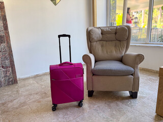 Pink suitcase standing next to a soft armchair in a hotel interior by a window. Travel, journey and...