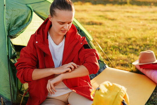 A displeased young woman sits by a tent, scratching her arm and looking annoyed at a bug bite.