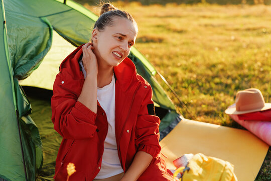 A distressed young woman sits by a tent, grimacing and scratching her neck due to an irritating bug bite.