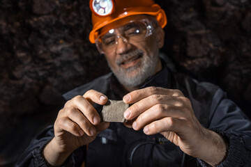 Hands of man in a hard hat with a flashlight examines rock, ore, rare earth metal. Geology,...