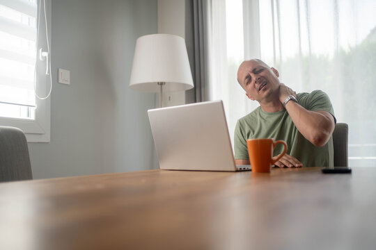 Mature man with neck pain working from home on laptop, experiencing discomfort and stiffness from prolonged sitting at computer
