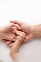 Parents&rsquo; hands gently holding a newborn&rsquo;s hand on white bedding. High-key lifestyle photo symbolizing love, safety, and early bonding&mdash;ideal for parenting, maternity, and healthcare use.