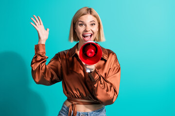 Obraz premium Youthful woman with blonde bob holding a red megaphone on turquoise background, radiating happiness and enthusiasm