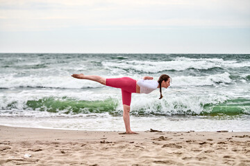 Young woman with brown hair practices yoga on sandy beach by baltic sea. Striking balance pose,...