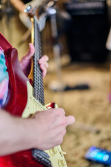 Close-up of young males hand playing red electric guitar in cozy studio. Warm lighting enhances vibrant atmosphere. Background shows blurred instruments and effects