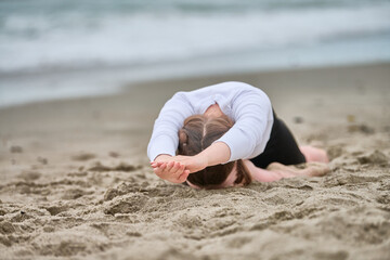 Young woman with brown hair practices yoga on baltic sea beach. Overcast sky casts soft light on sandy surface as she stretches, embodying calm and focus