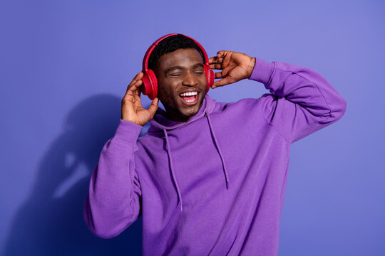 Young man enjoying music on red headphones while wearing a purple hoodie against a vibrant violet background