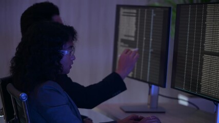 Professional cybersecurity analysts monitoring server data in dark office. Female developer pointing at code while collaborating with male colleague on urgent software project late at night. - Powered by Adobe