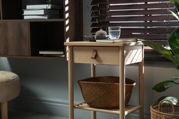 Wooden side table with glass of water, notebook, air reed diffuser and basket near window indoors