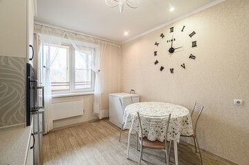 bright kitchen interior with light beige walls, a round table with floral tablecloth, metal chairs, a large clock on the wall, and a window with sheer curtains