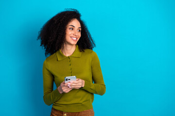 Charming young woman holding smartphone against vibrant blue background, showcasing casual style...