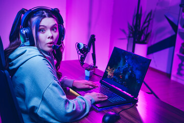 Joyful female gamer enjoying online streaming with headphones, laptop, and microphone in neon-lit...