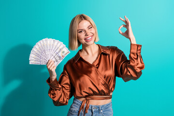 Elegant vibrant woman on a teal backdrop displaying cash and showing an okay gesture in stylish...