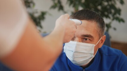 a man in a medical mask covid 19 has his temperature measured with a digital thermometer. coronavirus lockdown stay home social distance. temperature measurement mode during coronavirus