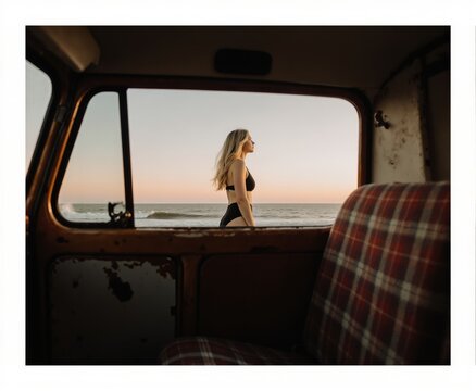 Fototapeta A woman in a swimsuit is seen through the window of a van, with the ocean and sunset in the background.