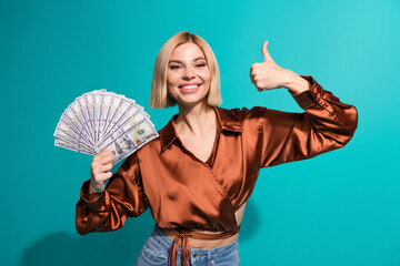 Happy young woman in a stylish satin blouse smiling with fan of money and giving thumbs up in a...