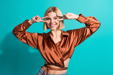 Young stylish woman smiling with peace sign in elegant satin blouse against a teal studio background