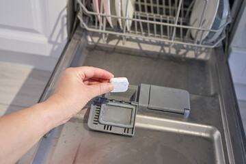 Hand placing dishwasher tablet in dishwasher compartment with open door and clean dishes visible.