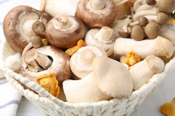 Different raw mushrooms in wicker basket on white table, closeup