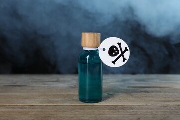 Bottle of poison with warning sign on wooden table against dark background with smoke