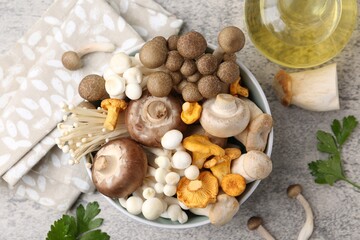 Different raw mushrooms in bowl, parsley and oil on grey textured table, flat lay