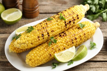 Tasty grilled corncobs with parsley and lime slices on wooden table, closeup