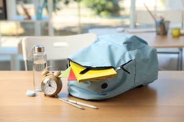 Backpack with different stationery, alarm clock and bottle of water on desk in classroom, closeup