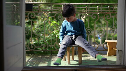 Young boy sitting on a small stool on the balcony, legs stretched out, enjoying a peaceful moment...
