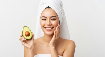 A smiling asian woman with a towel on her head holds half an avocado, her skin glowing after a spa treatment