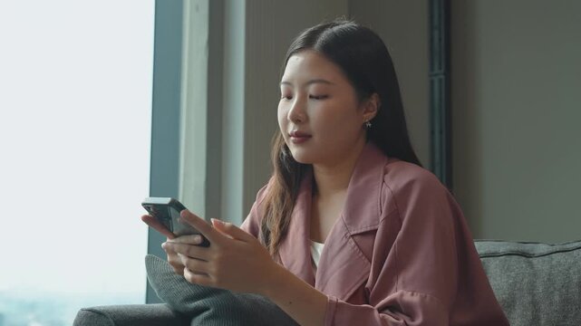 Happy young woman sitting on sofa holding phone smiling enjoying using mobile apps for shopping, having fun playing games, chatting on social media at office
