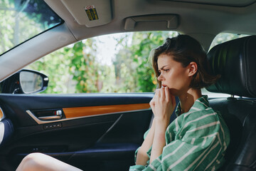 A thoughtful woman sits in a car interior, touching her lips with a worried, anxious expression as...
