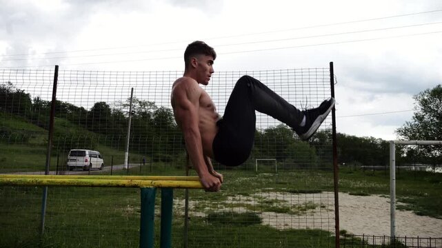 Strong guy training abdominal muscles on parallel bars at sports ground. Young shirtless sportsman exercising at nature. Sporty athlete working out at nature. Concept of active sportive lifestyle