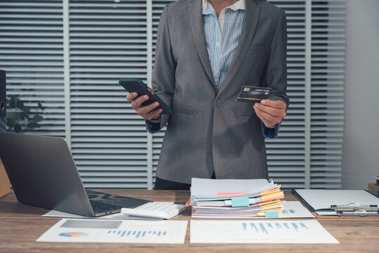 Digital Transaction: A professional stands at a desk in an office, smartphone in one hand and credit card in the other, symbolizing online business transactions. - Powered by Adobe