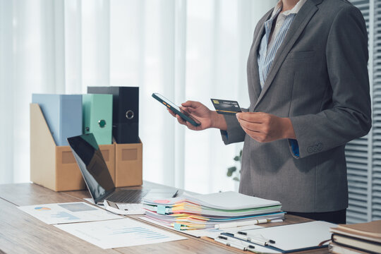 Businesswoman with credit card: a businesswoman uses a smartphone and holding a credit card standing behind a desk filled with office supplies. capturing business transaction.