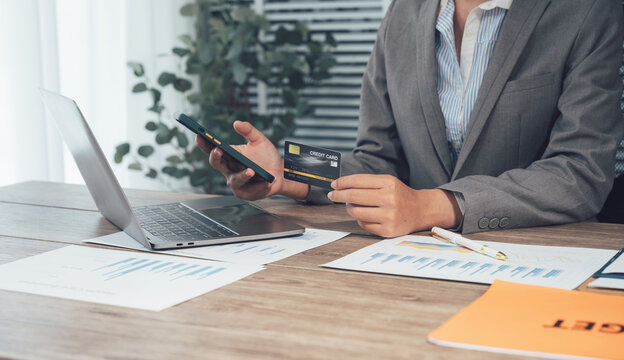 Business transaction with Credit Card: A professional, carefully manages an online business transaction, holding a credit card and smartphone near an open laptop and documents.