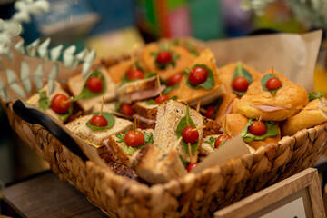 Rustic wicker basket holding ham and cheese sandwiches on wheat and white bread, garnished with cherry tomatoes and fresh basil