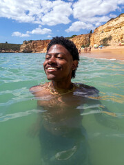 Young happy handsome African American man swims in idyllic ocean in Algarve Portugal Senhora da Rocha Beach Cliffs and blue sky background vacation tourism travel with copy space.
