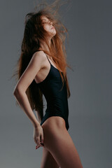 Young woman in black swimsuit posing with flowing hair on gray background, showcasing elegance and...