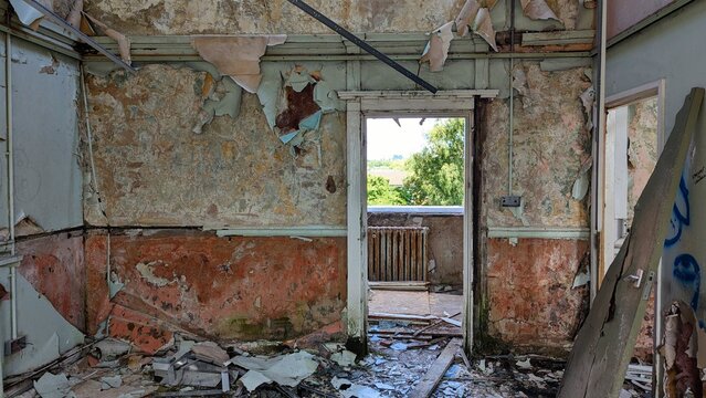 Crumbling plaster room walls, debris-covered floor, peeling wallpaper. Open balcony doorway falling apart. Derelict wrecked apartment contrast with nature outside. Urban decay interior disaster rubble