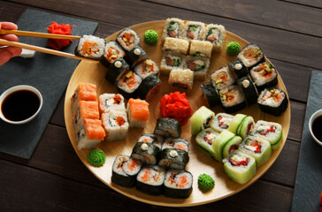 Japanese food restaurant, sushi maki gunkan roll plate or platter set. Hand take roll. Set for two with chopsticks, ginger, soy, wasabi. Sushi at rustic wood background and black stone. POV at black.